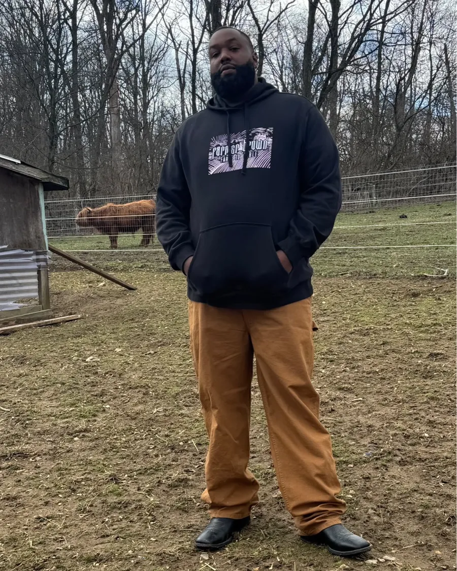 Grizz standing in the pasture in a Farmer Brown hoodie with a Highland cow grazing behind the fence
