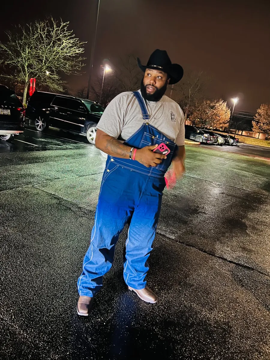 Grizz at night in a black cowboy hat, denim overalls and boots